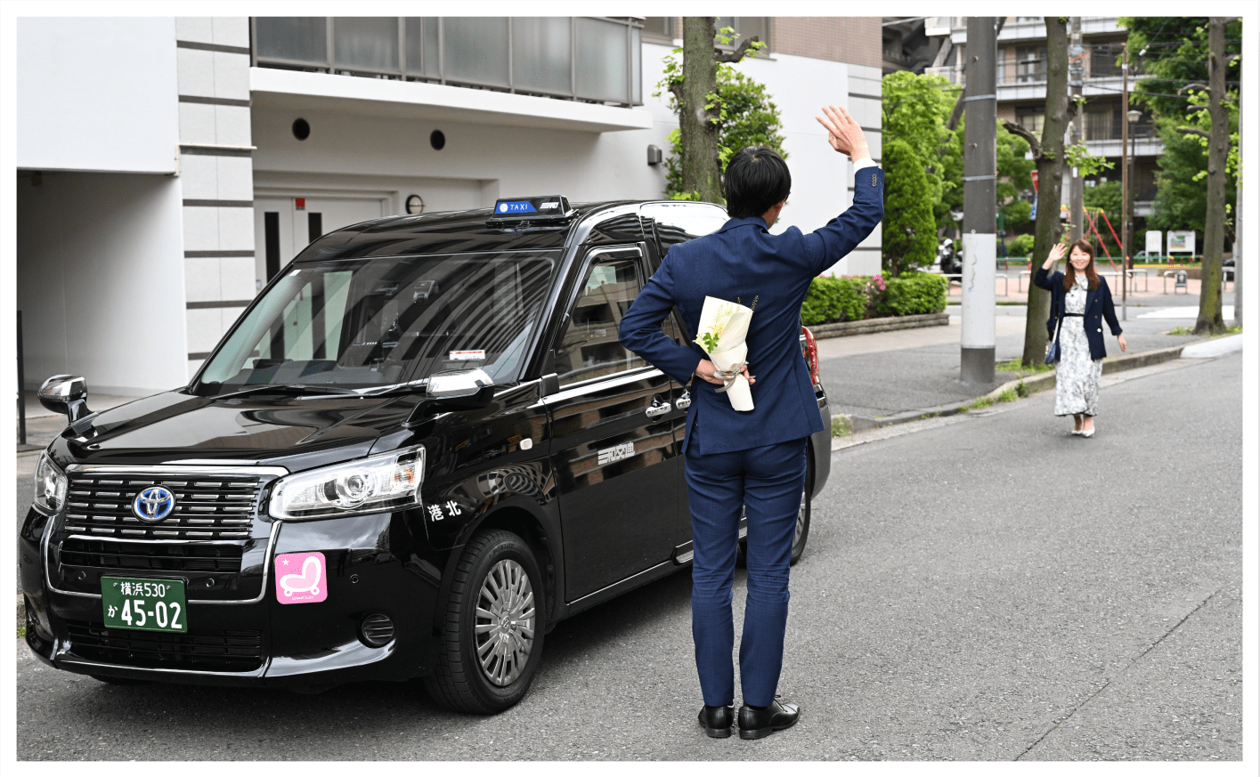 タクシー横で花束を後ろ手にお祝い相手を迎えるスーツの男性と手を振りながら近づいてくる女性