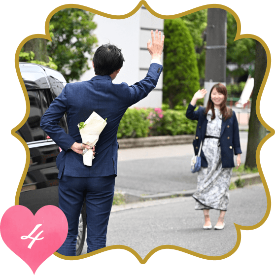 タクシー横で花束を後ろ手にお祝い相手を迎えるスーツの男性と手を振りながら近づいてくる女性