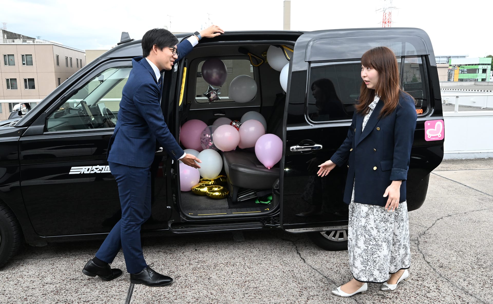 お祝い相手の女性をサプライズタクシーの後部座席にお迎えするスーツ姿の男性